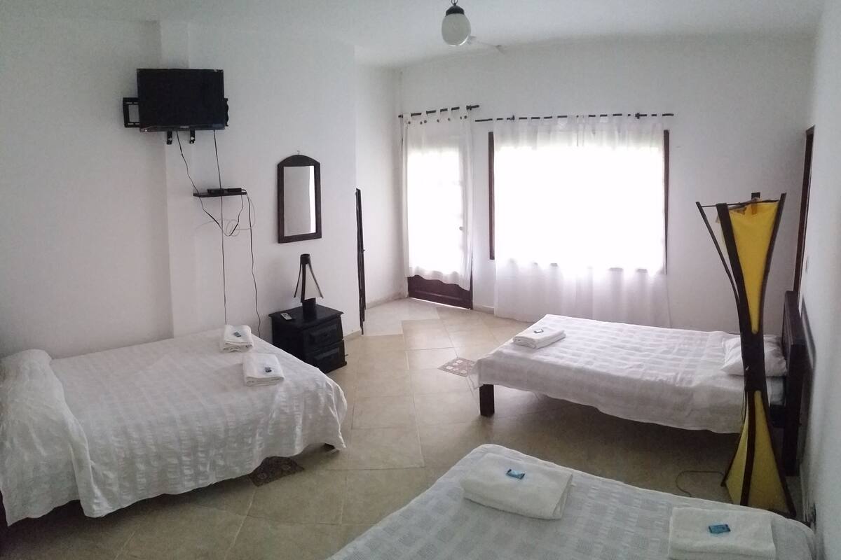 A room with two single beds, a flat-screen TV mounted on the wall, a mirror, and a window with sheer curtains.