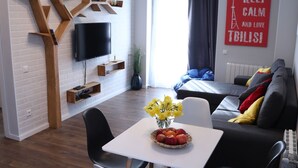 A modern living room with a white brick wall, a flat-screen TV, a dining table with chairs, and a sofa with cushions. - -30% off  Natalité Houzz Tbilisi (Tbilisi)