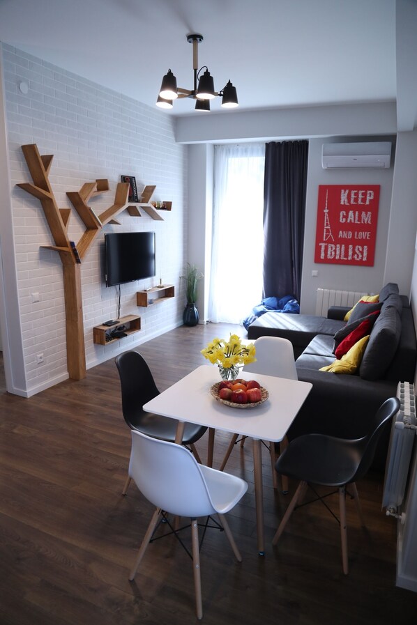 A modern living room with a white brick wall, a flat-screen TV, a dining table with chairs, and a sofa with cushions.