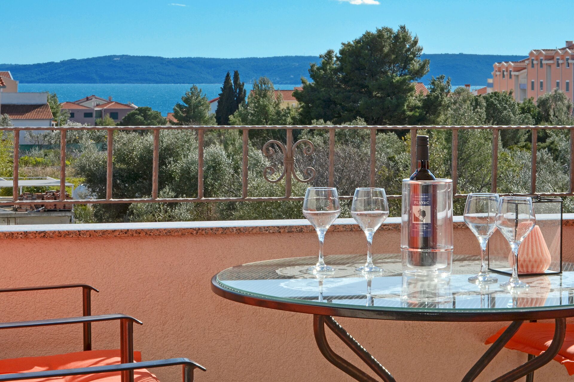 A glass table with wine glasses and a bottle on a balcony overlooking a body of water and hills.