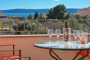 A glass table with wine glasses and a bottle on a balcony overlooking a body of water and hills.