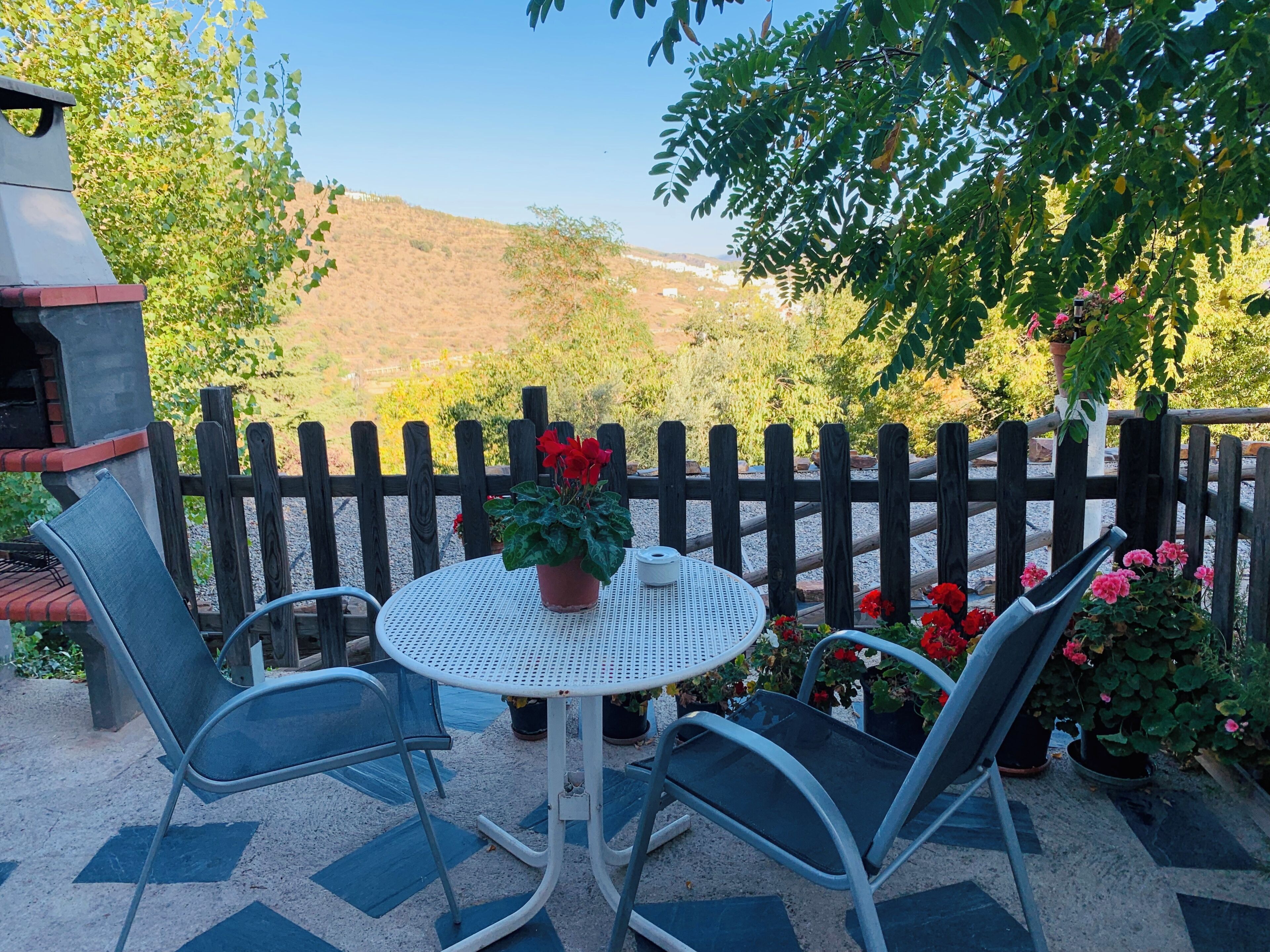 Een terras met een ronde tafel en twee stoelen, omringd door een houten hek, met uitzicht op een glooiend landschap op de achtergrond.