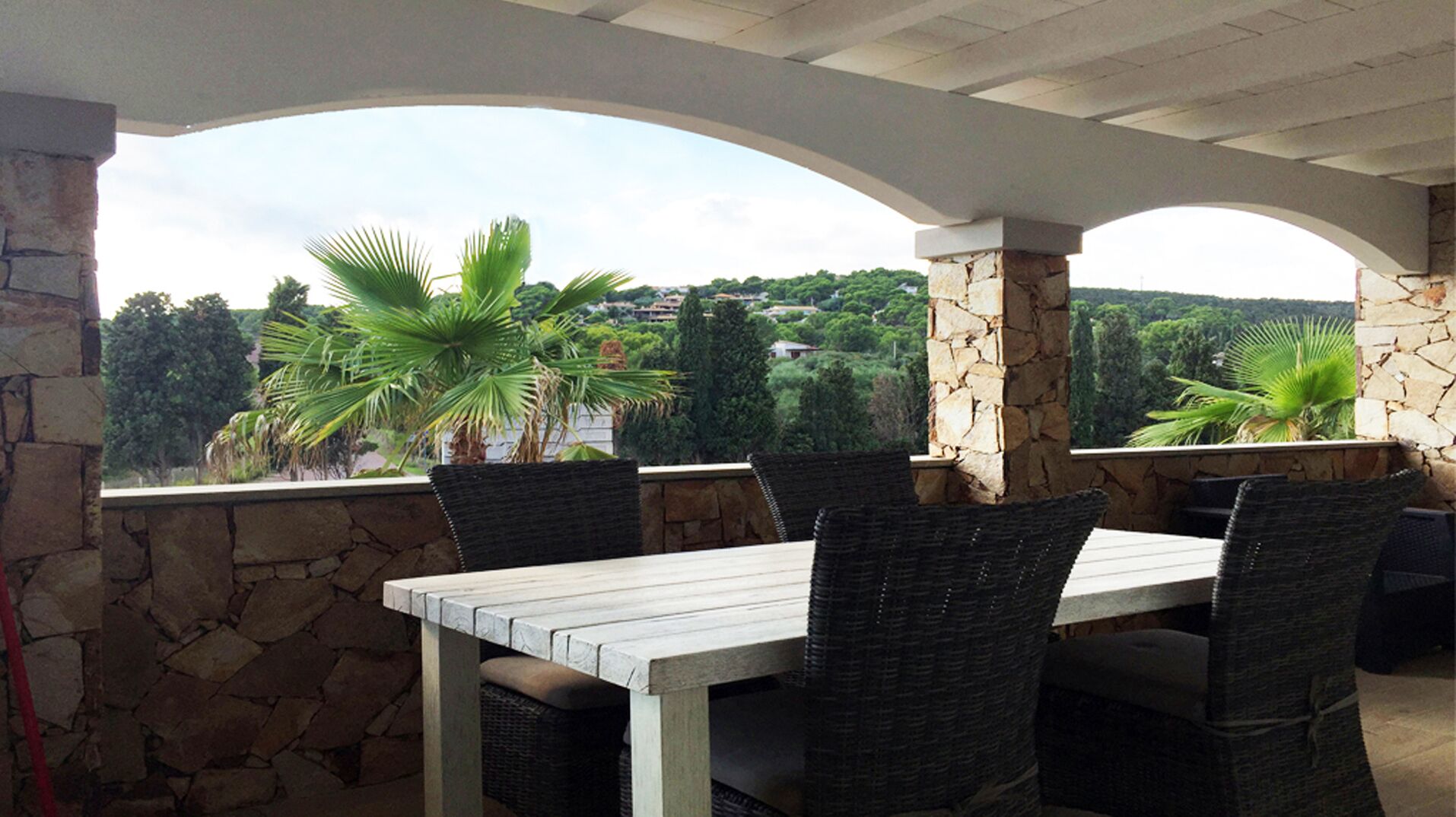 Une terrasse extérieure avec un mur en pierre, une table blanche et des chaises en osier, donnant sur un paysage luxuriant.