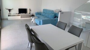 Un salón moderno con una mesa de comedor blanca, sillas grises, un sofá azul y un televisor sobre un mueble blanco.