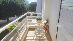 Un balcon avec une chaise et une table en métal peintes en blanc, un pot de fleurs et une vue sur les arbres et les bâtiments.