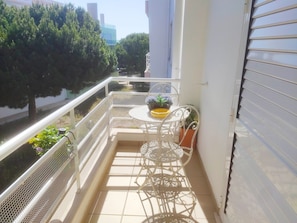 Varanda com cadeira e mesa de metal branco, um vaso de planta e vista para árvores e edifícios.