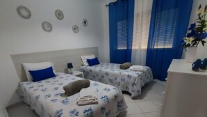 Une chambre d’hôtel avec deux lits, chacun agrémenté d’une couvre-lit à motifs floraux et de coussins bleus. On y trouve des décorations circulaires fixées au mur, une petite table de chevet avec une lampe, et un bureau sur lequel est posé un vase de fleurs.