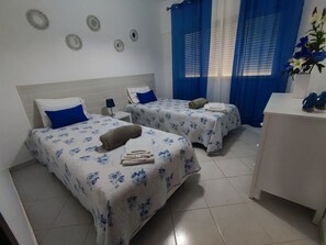 Quarto de hotel com duas camas, cada uma com colcha florida e almofadas azuis. Há decorações circulares na parede, uma pequena mesa de cabeceira com abajur e uma escrivaninha com um vaso de flores.