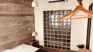 Een slaapkamer met een houten hoofdbord, een bed met een witte lakenset, een bedlamp, een houten kapstok, een raam met een ruitpatroon, en een airconditioning.