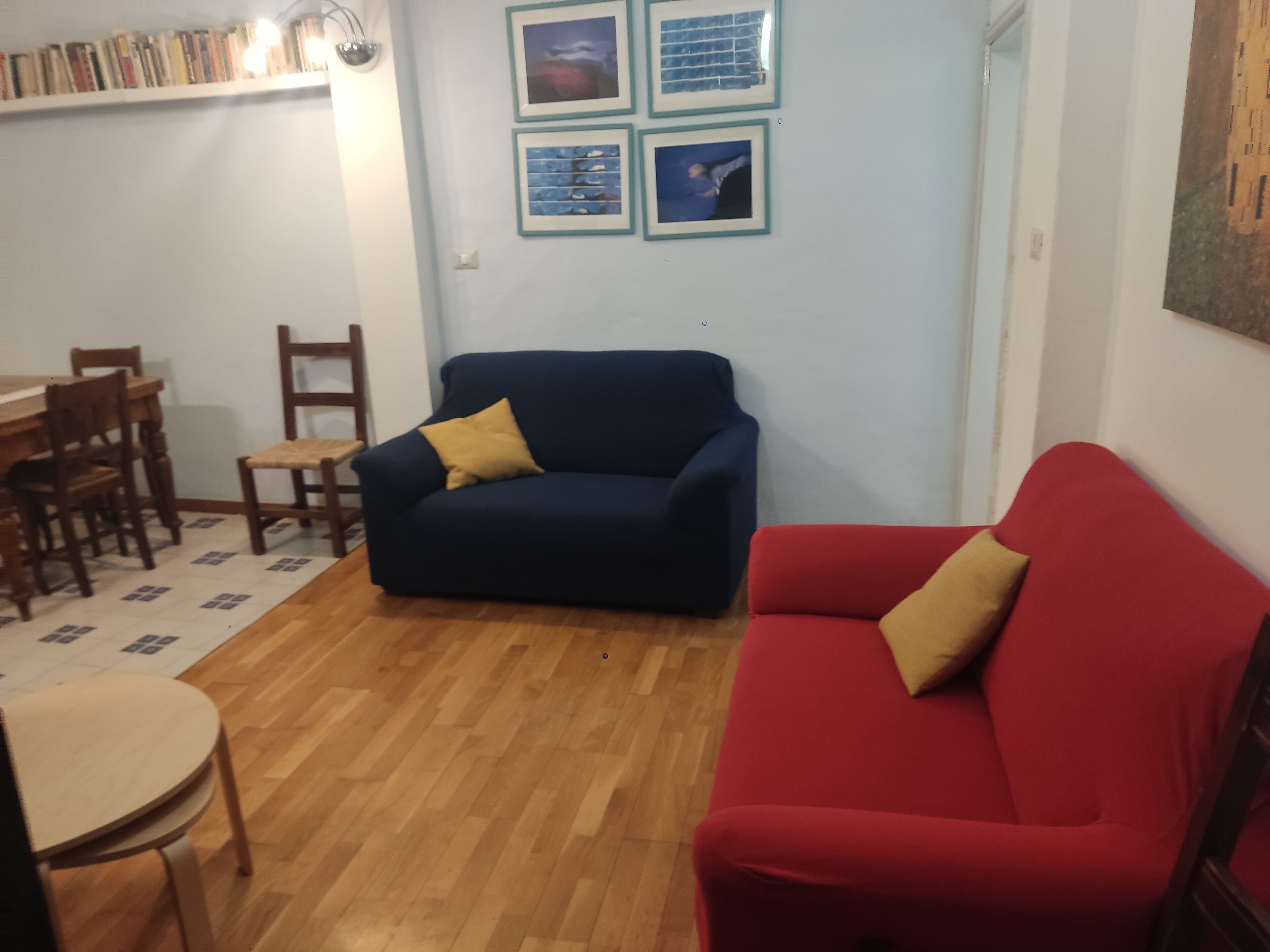 A living room with a blue sofa and a red sofa, wooden chairs, a small round table, and a bookshelf filled with books.