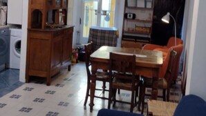Una sala de estar con un sofá azul, una mesa de comedor de madera y sillas. Se observa una zona de cocina con lavadora y un armario de madera.