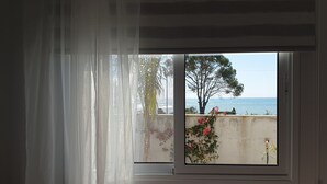 Ein Fenster mit Meerblick und einem Baum, teilweise zurückgezogene Vorhänge und eine Pflanze auf dem Fensterbrett.