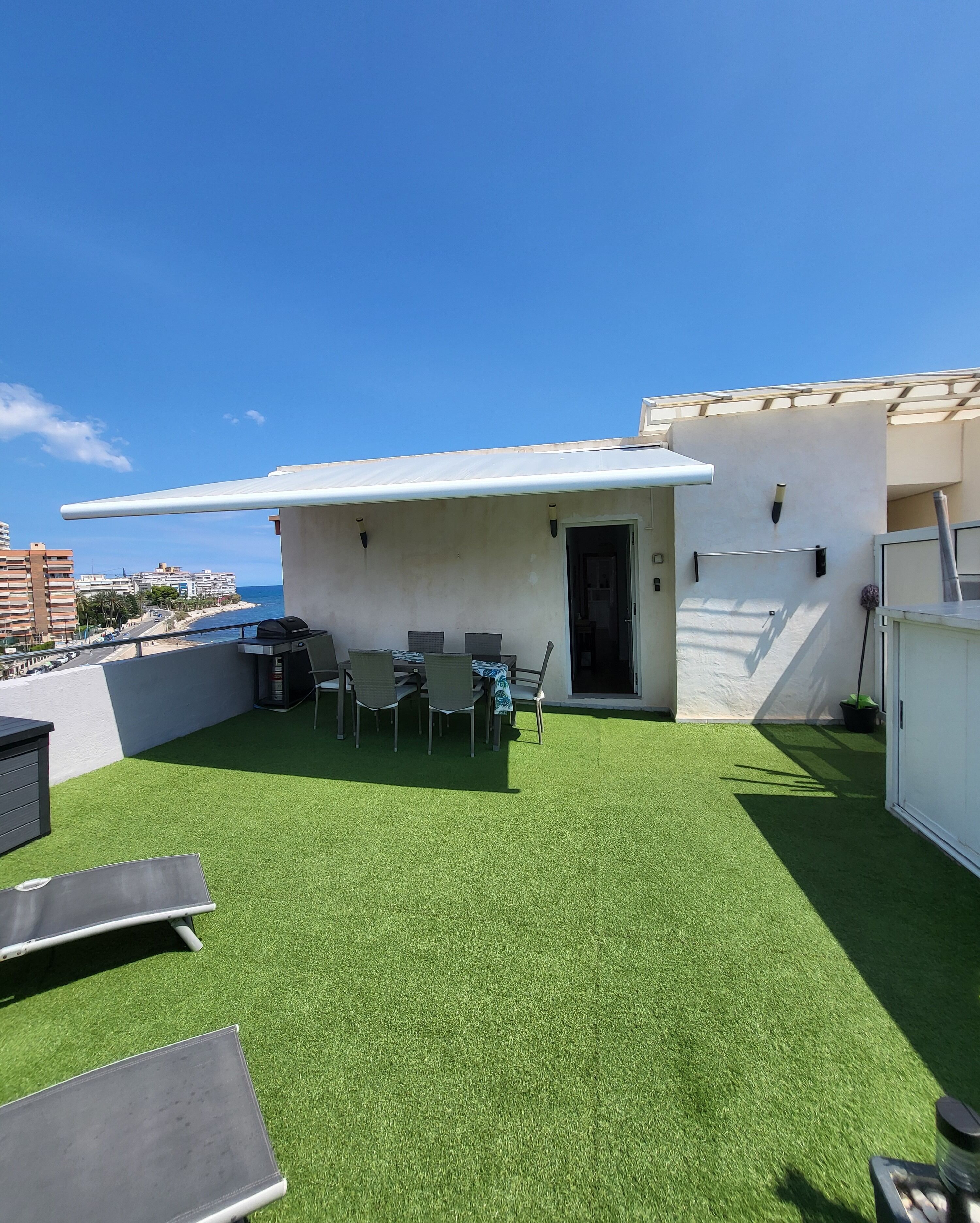 Terraço no telhado com grama sintética, uma mesa de jantar com cadeiras, churrasqueira e vista para o mar.