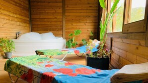 Una cabaña de madera con dos camillas de masaje cubiertas con sábanas florales coloridas, un jacuzzi y plantas en macetas.