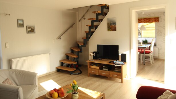 Uma sala de estar moderna com escada de madeira, televisão de tela plana e uma área de jantar visível por uma porta aberta.