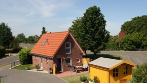 Uma casa de dois andares com telhado vermelho, uma pequena construção amarela anexa e um caminho de acesso pavimentado.
