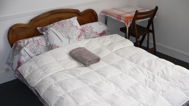 A neatly made bed with a white quilt and floral pillows, a wooden headboard, and a small table with a plaid tablecloth and a chair. - Large detached comfortable room (Le Mesnil-Esnard)