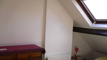 Une chambre avec un plafond en pente, une fenêtre, un radiateur, une commode en bois, une petite table avec une lampe et un lit recouvert d’une couverture jaune.