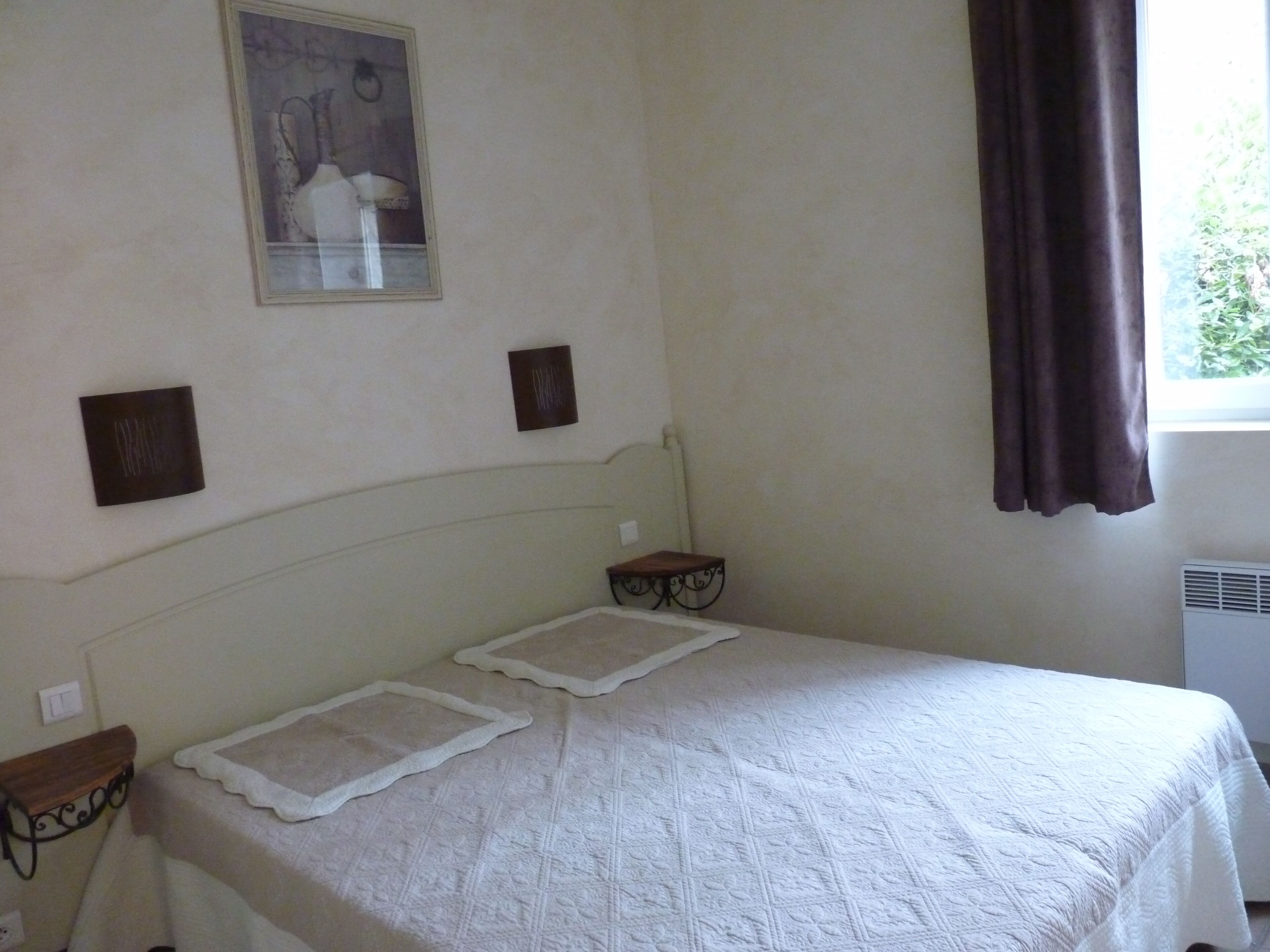 Une chambre à coucher comprenant un lit, une table de chevet, une étagère fixée au mur, un tableau encadré et une fenêtre avec des rideaux.