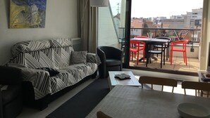 Een moderne woonkamer met een bank, een eettafel en stoelen. Er is een balkon met rode stoelen en een tafel, en een uitzicht op de stad door het raam.