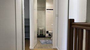 Een gang met houten vloer die leidt naar een badkamer met douche, toilet en een klein vloerkleed.