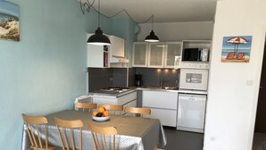 Een kleine keuken met witte kastjes, een magnetron en een vaatwasser. Er staat een eettafel met vier stoelen en een schaal met fruit.