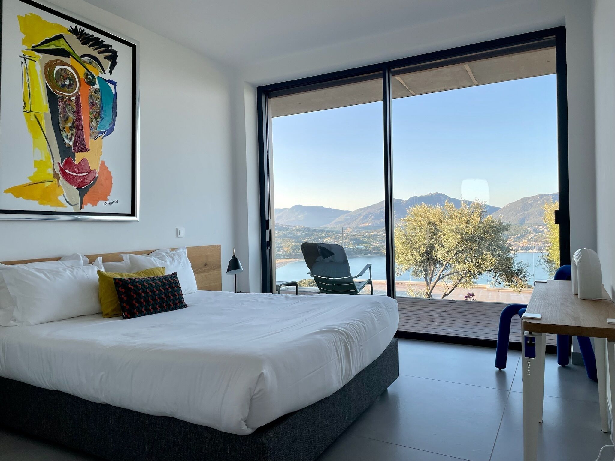 Habitación de hotel moderna con una cama grande, un escritorio y una silla. La habitación tiene un ventanal amplio que ofrece vistas a un lago y montañas.