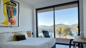 A modern hotel room with a large bed, a desk, and a chair. The room has a large window offering a view of a lake and mountains. - Villa Lourada architect house, swimming pool, spa 1.5 km from beautiful beaches  (Olmeto)