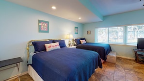 A room with two beds, a window with blinds, a desk with a chair, and a television.