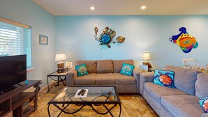 A living room with a sofa, two side tables with lamps, a glass coffee table, and a flat-screen TV. - Shared Pool, Walk Path to Beach, Golf Cart Access (Port Aransas)