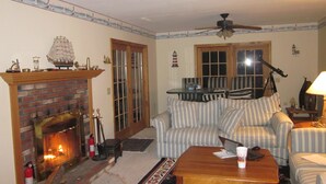 A cozy living room with a fireplace, striped sofa, and a ceiling fan. - Luxury Lakefront Living in Lake Sebago Area (Bridgton)