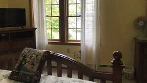 A bedroom with a large window, a bed, a chair, a dresser, a television, and a fan. - Luxury Lakefront Living in Lake Sebago Area (Bridgton)
