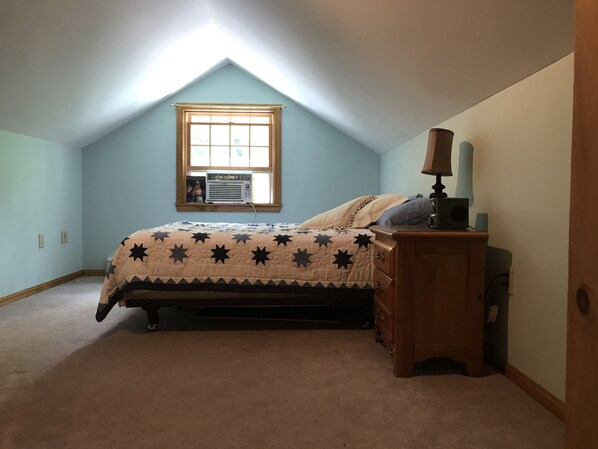 A bedroom with a sloped ceiling, a window with an air conditioner, a bed with a star-patterned quilt, a nightstand with a lamp, and a dresser.