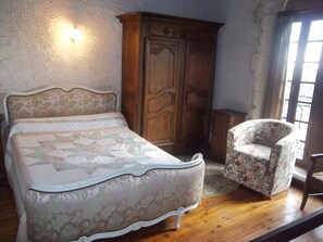 Een slaapkamer met een bed, een houten garderobe, een fauteuil met bloemenprint en een raam met natuurlijk licht.