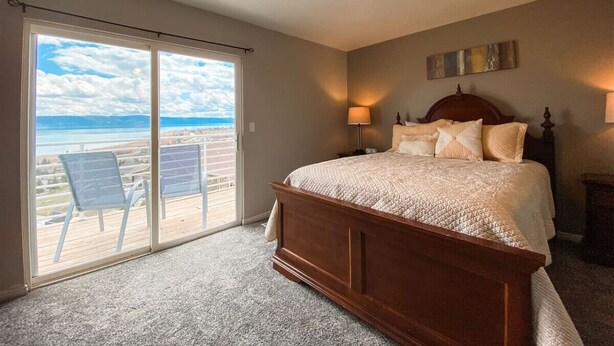 A bedroom with a large bed, a ceiling fan, a view of the beach through a sliding door, and a nightstand with a lamp.