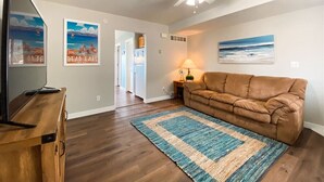 A living room with a brown leather sofa, a wooden coffee table, a TV, and a ceiling fan. There are framed pictures on the walls and a rug on the floor.