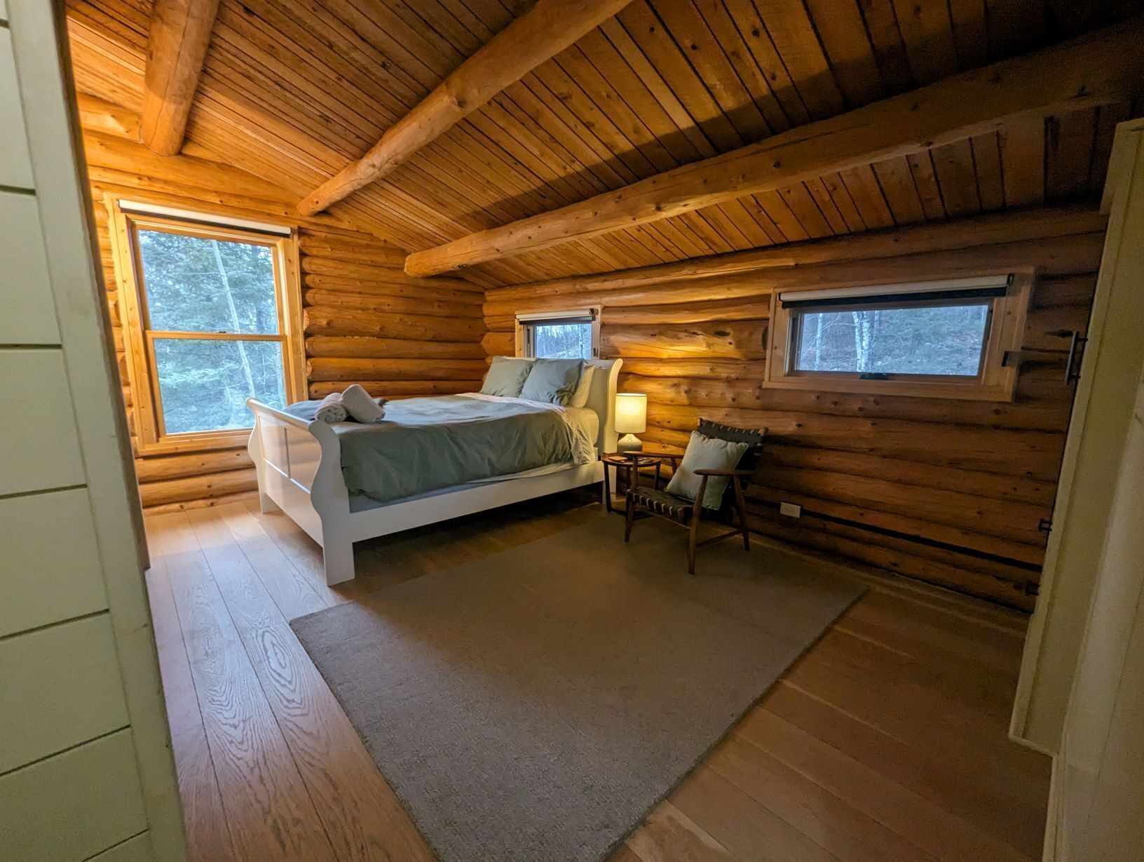Un dormitorio tipo cabaña de madera con una cama, dos ventanas, una mesita de noche y una silla.