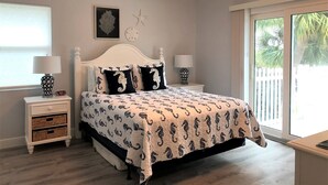 A bedroom with a bed, two bedside tables, a ceiling fan, a clock, and a window with a view of palm trees. - Beach Cottage, Steps to Beach. Updated and Great Deal for Money. Pet Friendly. (Fort Pierce)