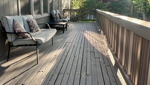 Una terraza de madera con sofá, sillas, barandilla y vistas a árboles.