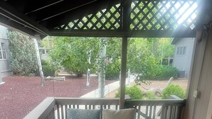 A covered patio with a glass-top table and two chairs, a wooden deck, and a view of a garden and house in the background. - Amazing 5 Star Home, Amenities Galore, AC, no pet fee, 7 miles to NAU (Flagstaff)