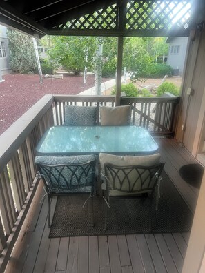 Une terrasse couverte avec une table à plateau de verre et deux chaises, une terrasse en bois et une vue sur un jardin et une maison en arrière-plan.
