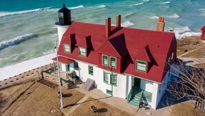 A coastal house with a red roof and white walls, situated near the ocean with waves crashing onto the shore. - Year round Crystal Lake cottage, newly remodelled 4br/1ba, w/sandy beach! (Frankfort)