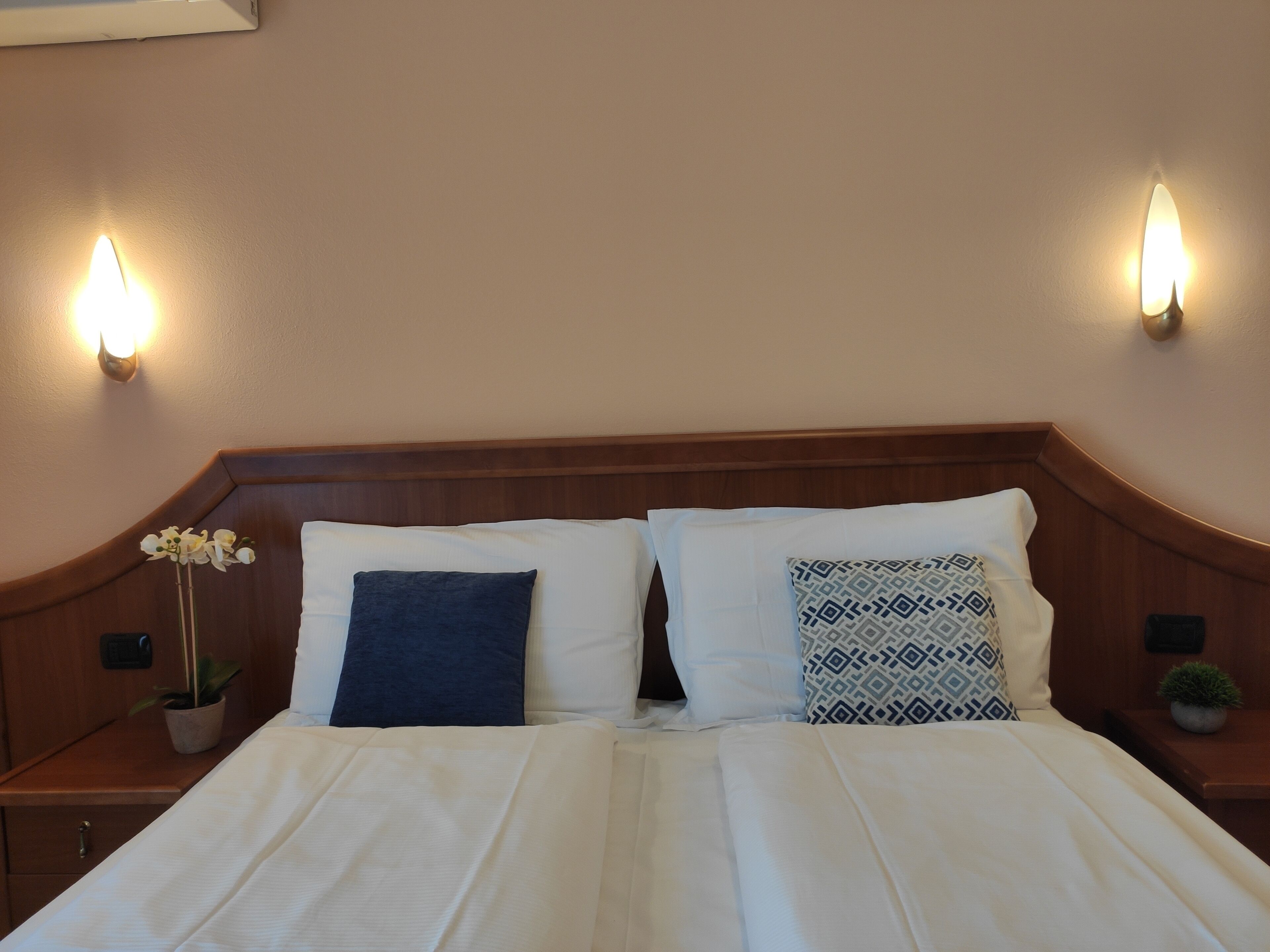 Ein Doppelbett mit weißen Laken, zwei Kissen, ein Nachttisch mit einer Topfpflanze und Wandleuchten.