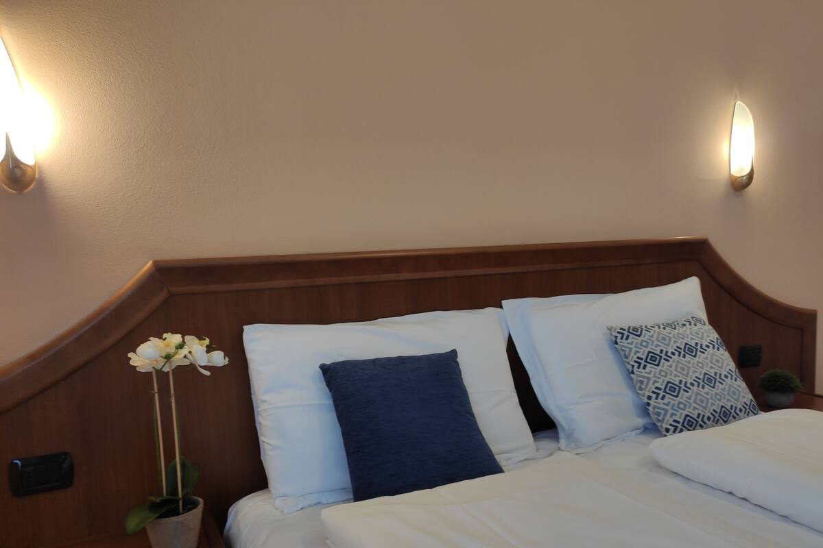 Habitación de hotel con cabecera de madera, cama con ropa blanca, una orquídea en maceta y apliques de pared.