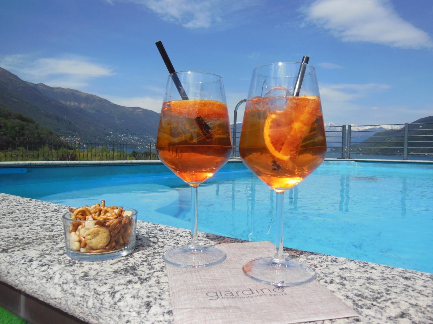 Two glasses of a beverage with an orange slice, a glass of nuts, and a pool with a mountainous backdrop.