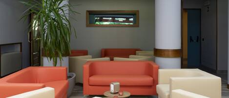 A lobby with a red sofa, beige armchairs, a potted plant, and a small table with a lamp.