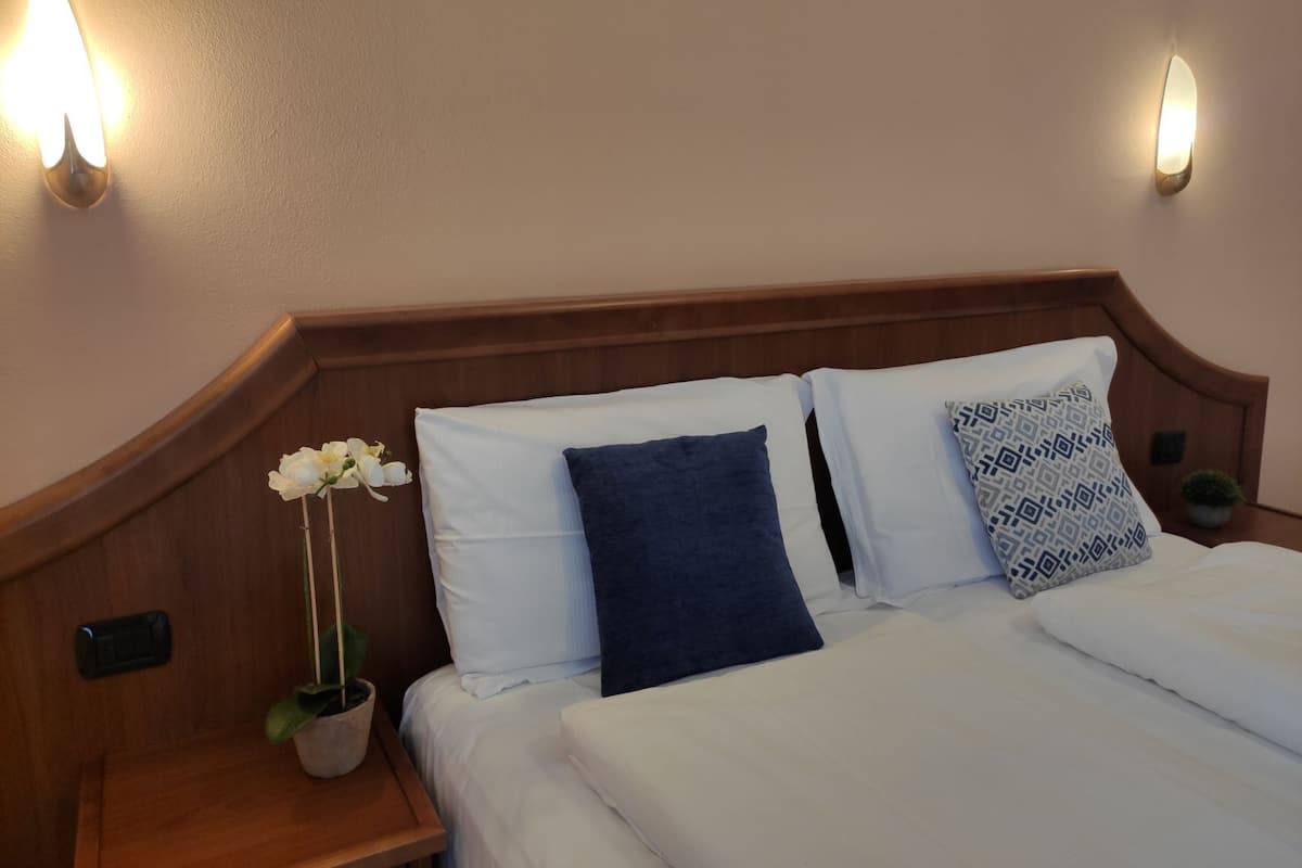 Habitación de hotel con cabecera de madera, dos almohadas blancas, una almohada azul y una almohada estampada. Hay una planta en maceta sobre la mesita de noche.