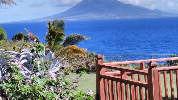 Vista costera con montaña al fondo, cerca de una valla de madera y vegetación tropical.