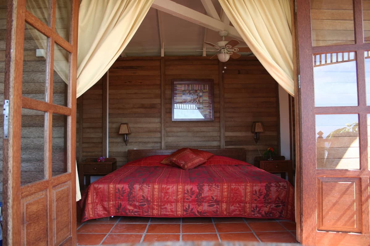 A wooden room with a bed, curtains, a ceiling fan, and a window.