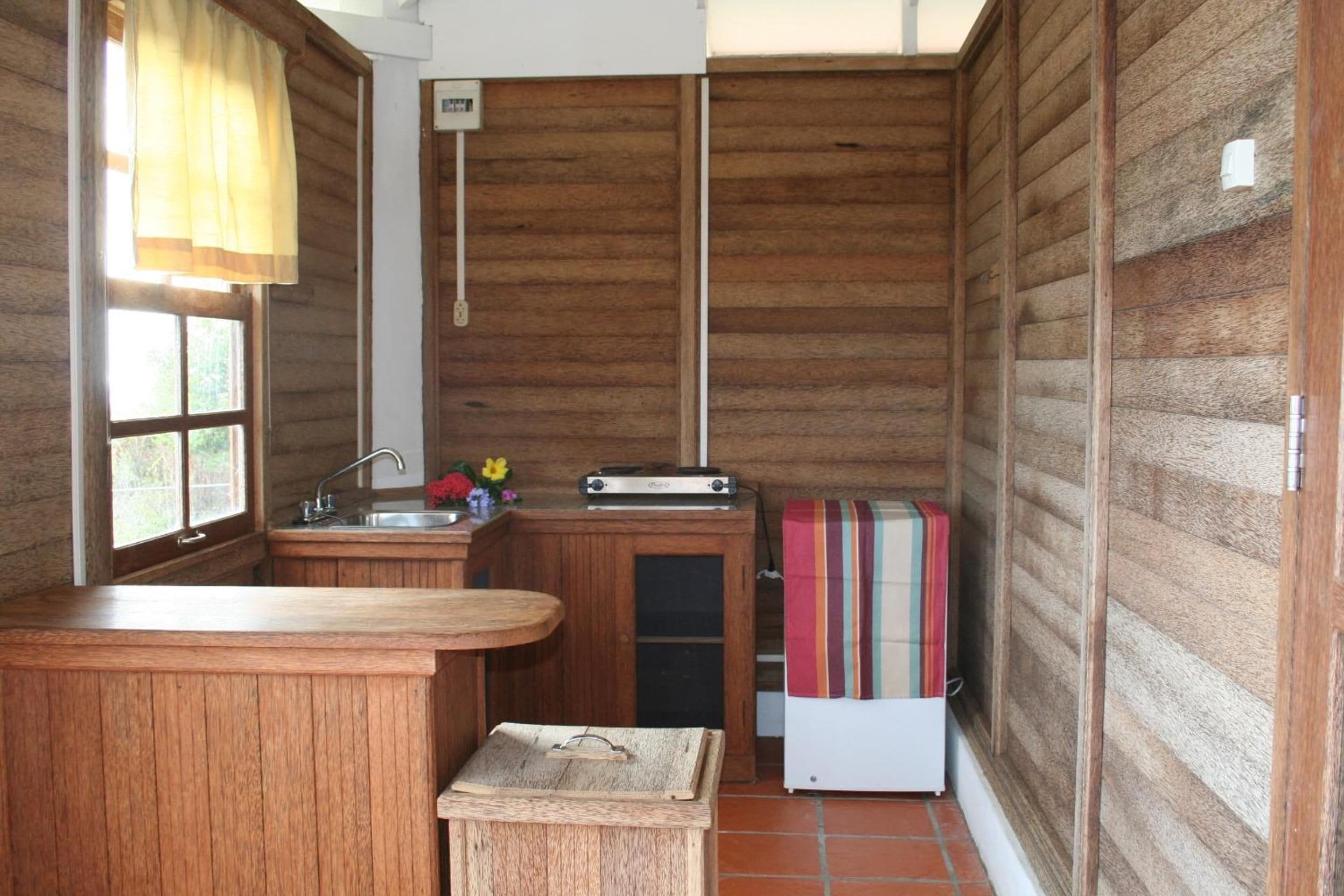 A compact kitchen with wooden walls, a sink, a small refrigerator, and a window with curtains.
